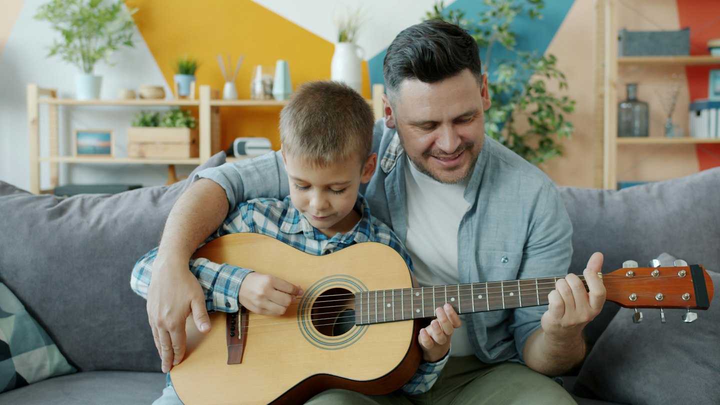 Man teaches boy to play guitar on a sofa.