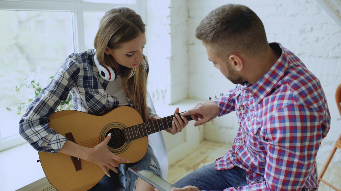 A man points to the guitar fretboard while teaching a woman.